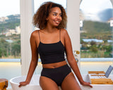 Woman in black period underwear and top sitting on a white bathtub with a scenic view outside.